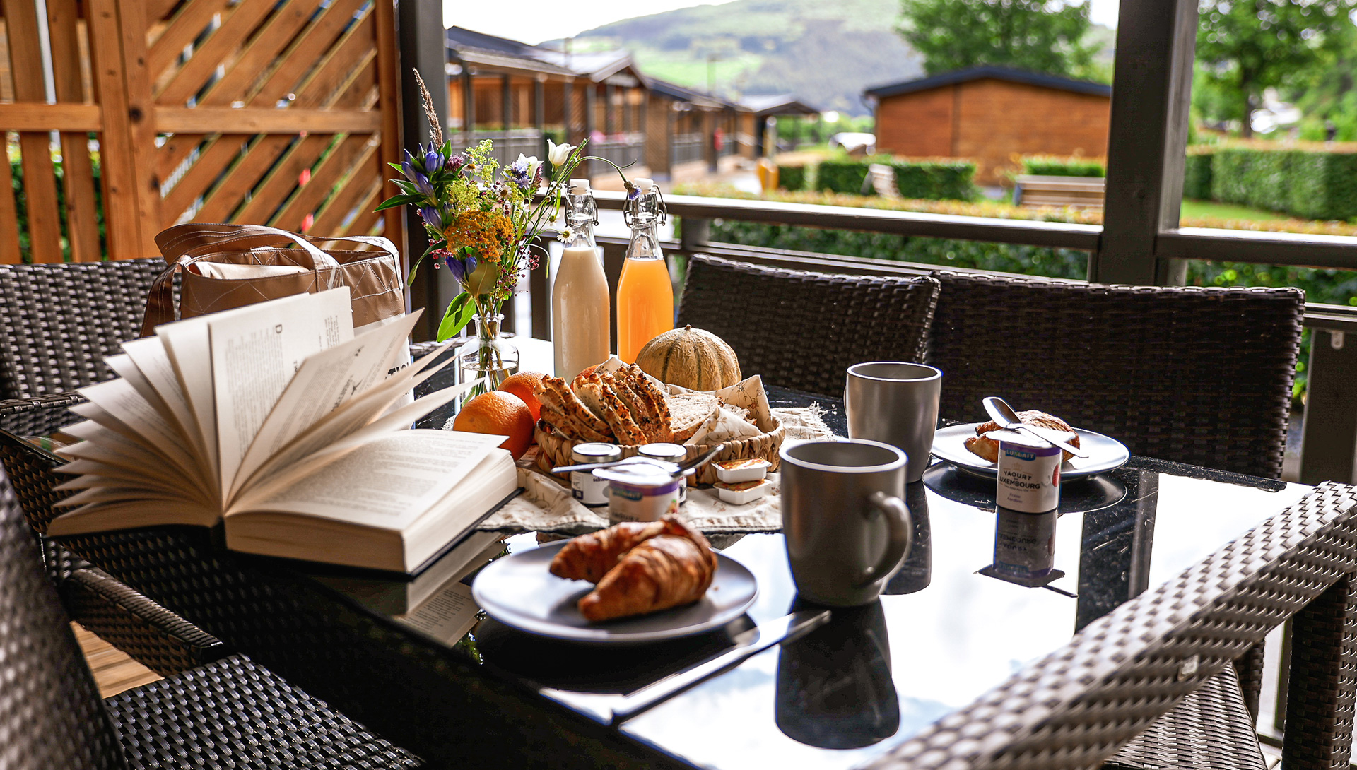 Frühstück auf der Terrasse eines Chalets mit Croissants, Brot, Marmelade, Saft und Kaffee – Blick auf weitere Chalets des Camping Kaul